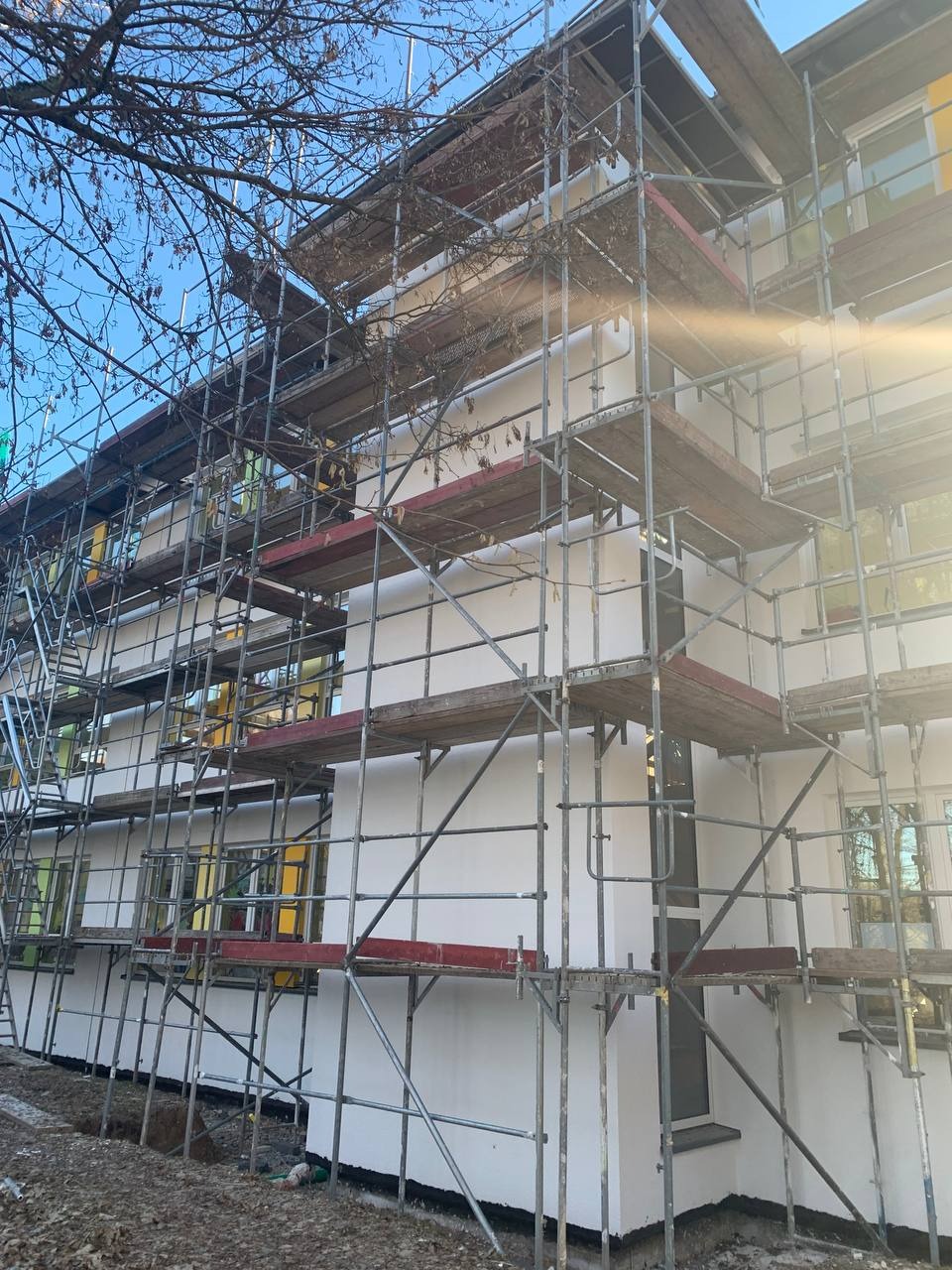Budynek wielopiętrowy w trakcie prac elewacyjnych, otoczony rusztowaniami, z widocznymi oknami z żółtymi ramami i świeżo pomalowaną białą elewacją.