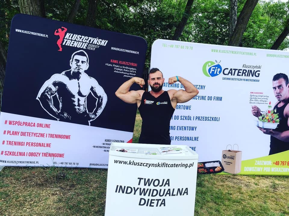 Mężczyzna z brodą, prezentujący muskulaturę na tle banerów reklamowych Kluszczyński Trenuje i Fit Catering, z ekspozytorem 'Twoja indywidualna dieta' i pudełkiem cateringu dietetycznego.