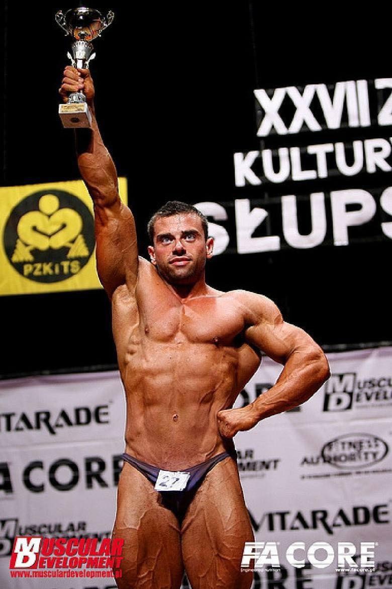 Mężczyzna kulturysta w stroju startowym, trzymający trofeum nad głową na scenie z banerami sponsorskimi w tle. Widoczny numer startowy '27'.