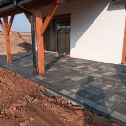 Nowo ułożona, szara kostka brukowa na tarasie domu z drewnianą konstrukcją zadaszenia, widoczny fragment ziemi przed tarasem.