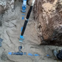 Wykonane przyłącze wodociągowe w wykopie, rury niebieskie i czarne zasypane piaskiem, widoczny fragment buta pracownika.