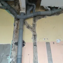 Szare rury kanalizacyjne z licznymi kolankami, zamontowane w ścianie z widocznymi śladami remontu i fragmentami starej farby w odcieniach żółci i różu, z fragmentem sufitu z wyciętym otworem.