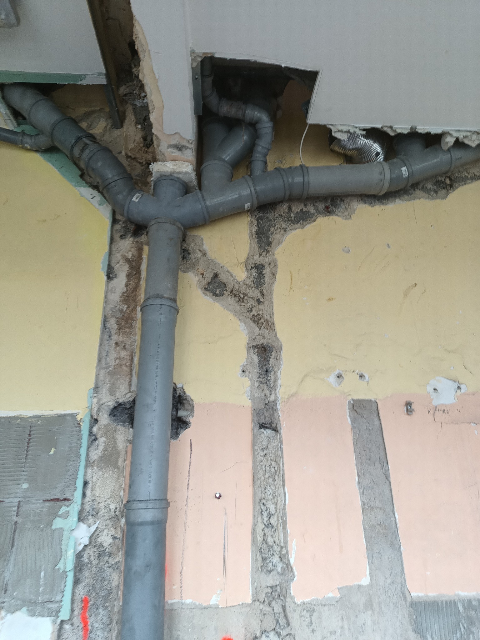 Szare rury kanalizacyjne z licznymi kolankami, zamontowane w ścianie z widocznymi śladami remontu i fragmentami starej farby w odcieniach żółci i różu, z fragmentem sufitu z wyciętym otworem.