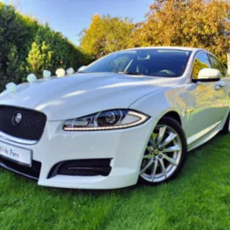 Biały Jaguar udekorowany białymi kwiatami na masce i tablicą 'Młoda Para', stojący na trawniku przed domem w słoneczny dzień.