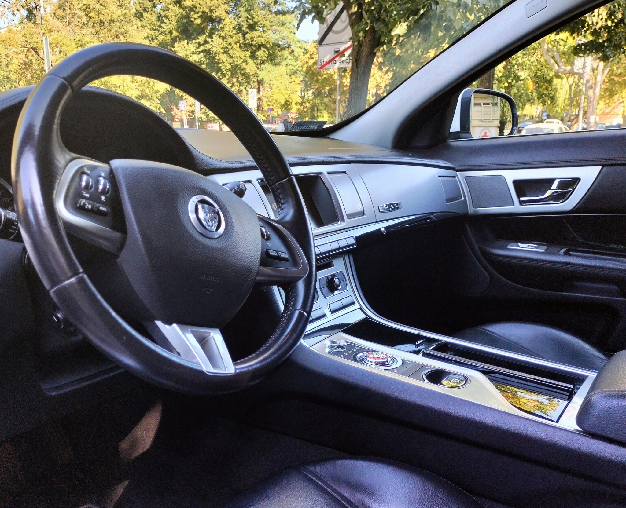 Wnętrze luksusowego samochodu Jaguar z widoczną kierownicą, deską rozdzielczą z chromowanymi elementami i skórzanymi siedzeniami, z zewnątrz widoczne drzewa.