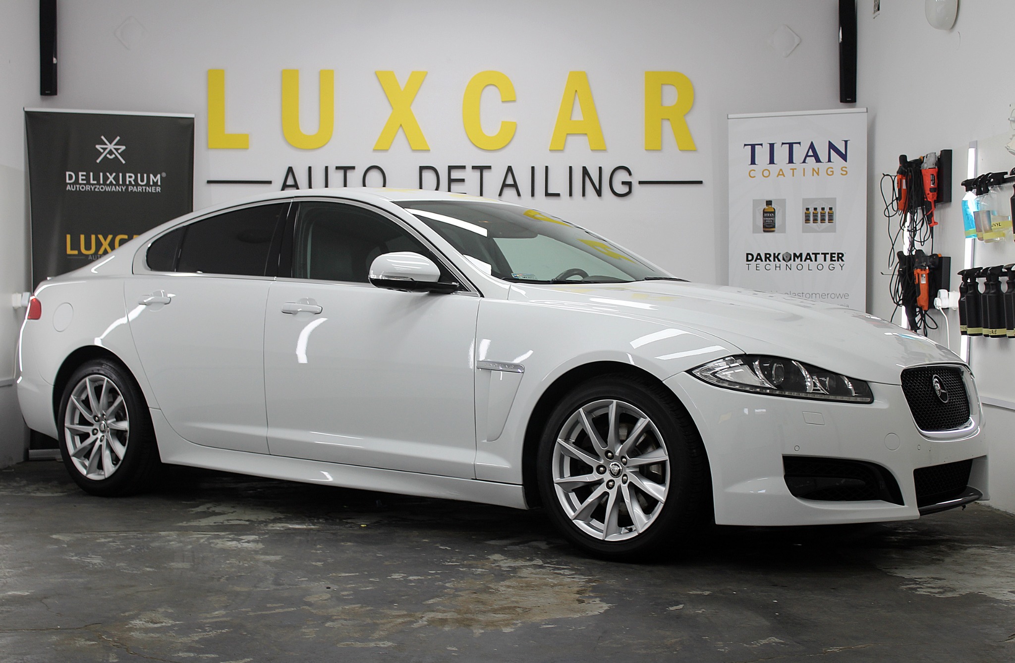 Biały Jaguar XF po detailingu w studiu Luxcar, widoczne logo Delixirum i Titan Coatings na ścianie, profesjonalne oświetlenie i sprzęt do pielęgnacji aut.