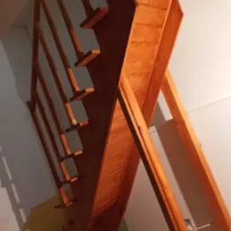 Drewniane schody strychowe z barierką, widoczne od dołu, z elementami dekoracyjnymi na stopniach i podporach, pomalowane na ciepły, brązowy kolor.