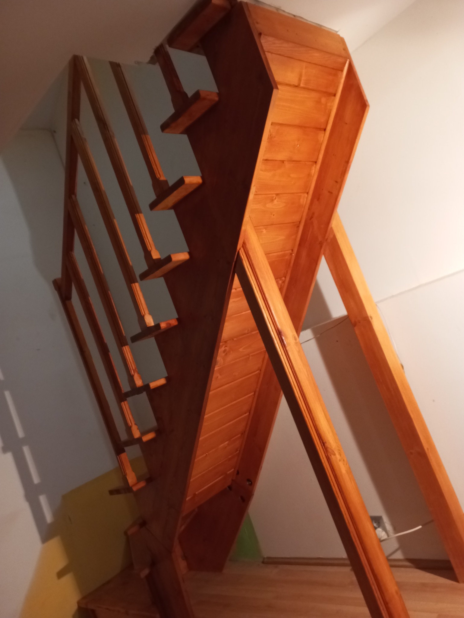 Drewniane schody strychowe z barierką, widoczne od dołu, z elementami dekoracyjnymi na stopniach i podporach, pomalowane na ciepły, brązowy kolor.