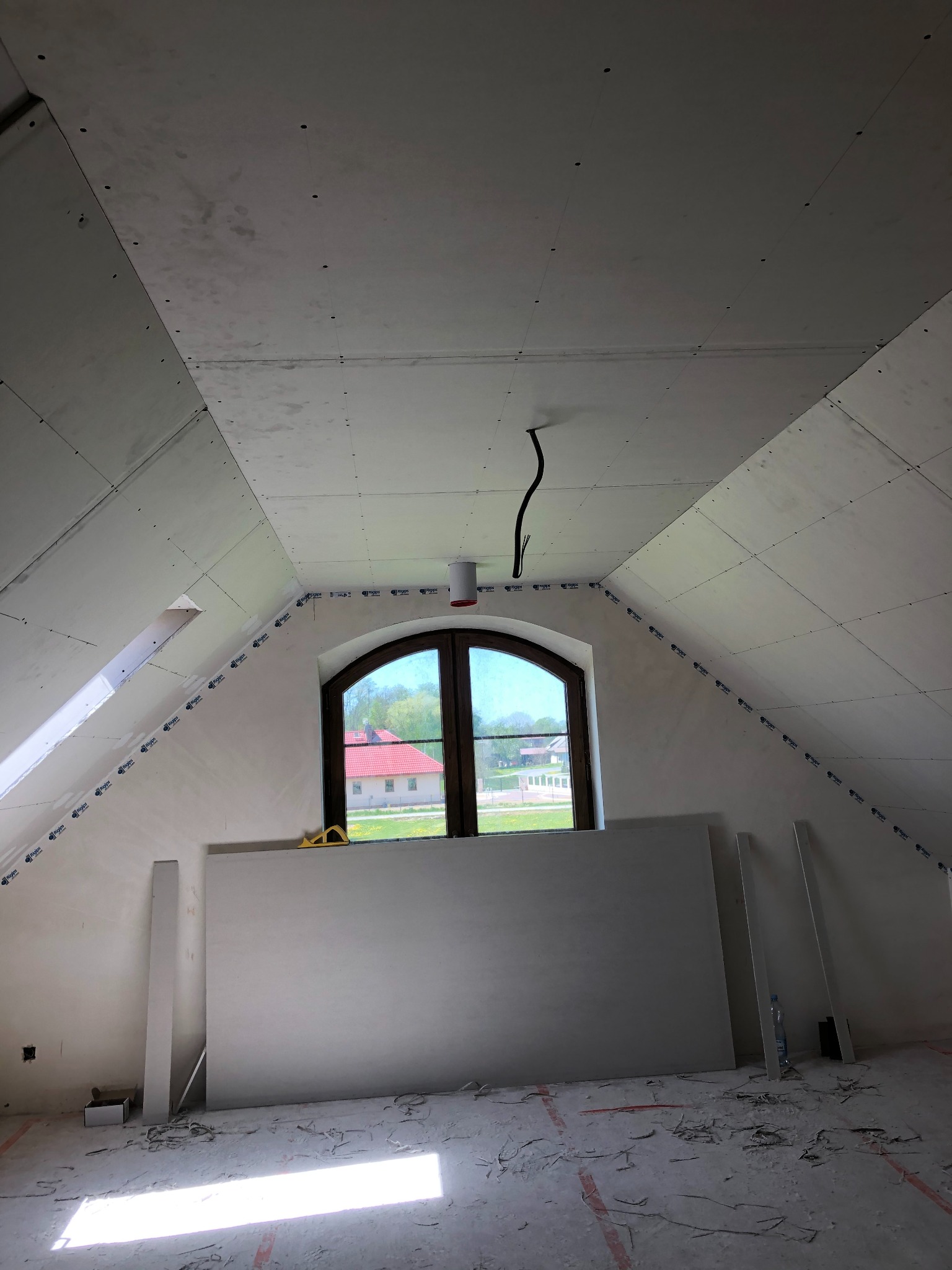 Poddasze z oknem w trakcie montażu płyt gipsowo-kartonowych na skośnych ścianach i suficie. Widoczne łączenia płyt i wystające przewody elektryczne.
