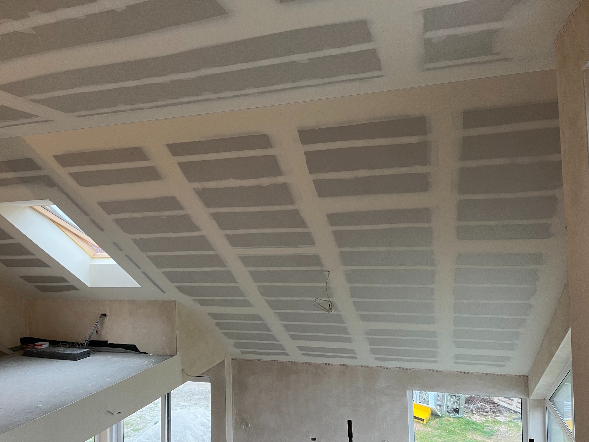 Skośny sufit poddasza z płyt kartonowo-gipsowych, widoczne łączenia płyt, przygotowany do dalszych prac wykończeniowych. Okno dachowe w tle.