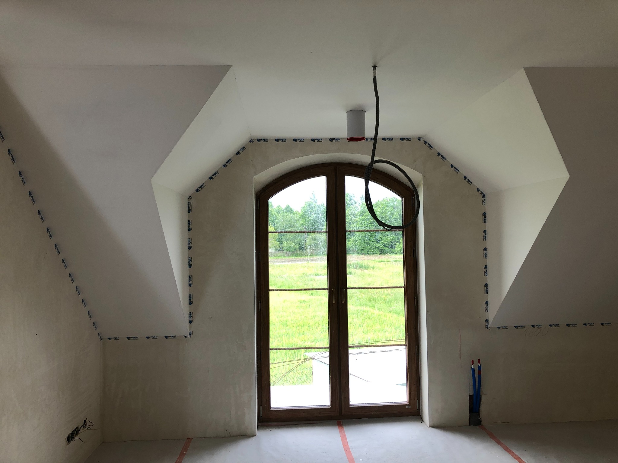 Poddasze po szpachlowaniu, przygotowane do malowania. Widoczne okno z drewnianą ramą, zabezpieczone ściany taśmą malarską i wystające rury w rogu.