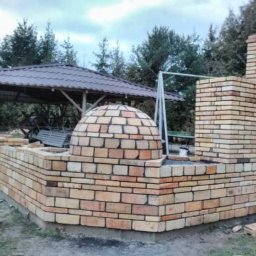 KP Project - Murowana konstrukcja z cegły klinkierowej w ogrodzie, z kopułą i kominem, obok altana z metalowym dachem, widoczne drzewa w tle.