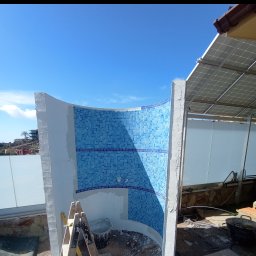 KP Project - Niedokończona, półokrągła ściana prysznicowa z niebieskiej mozaiki, z widocznymi śladami zaprawy, drabina i wiadro na placu budowy, jasne niebo w tle.