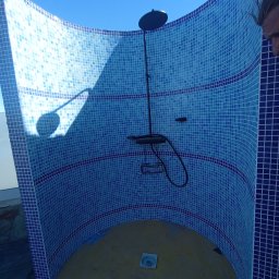 KP Project - Niedokończona kabina prysznicowa z zaokrąglonymi ścianami wyłożonymi mozaiką w odcieniach błękitu, z widoczną armaturą i odpływem w podłodze, w trakcie prac wykończeniowych.