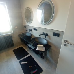 KP Project - Nowoczesna łazienka z dwoma czarnymi umywalkami nablatowymi, owalnymi lustrami z podświetleniem LED, szarą płytką ścienną i podłogową, czarnymi bateriami oraz grzejnikiem żeberkowym; na podłodze...