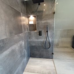 KP Project - Nowoczesna kabina prysznicowa z szarymi płytkami imitującymi beton, czarna deszczownica i bateria, szklana ściana kabiny, oświetlona półka w ścianie.