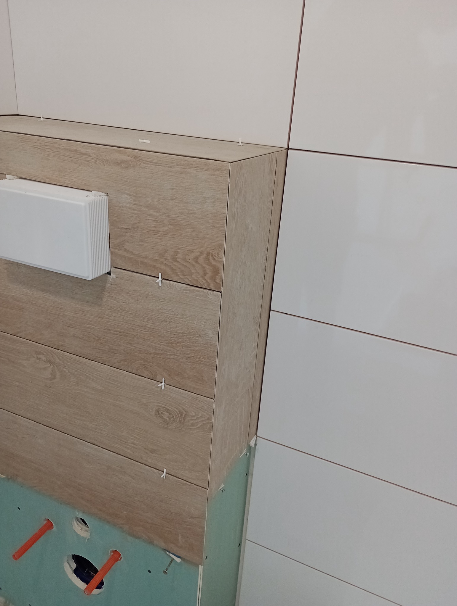Fragment łazienki w trakcie wykańczania: płytki imitujące drewno na zabudowie stelaża WC, białe, poziome płytki na ścianie, widoczne krzyżyki dystansowe, instalacja wodna w ścianie.