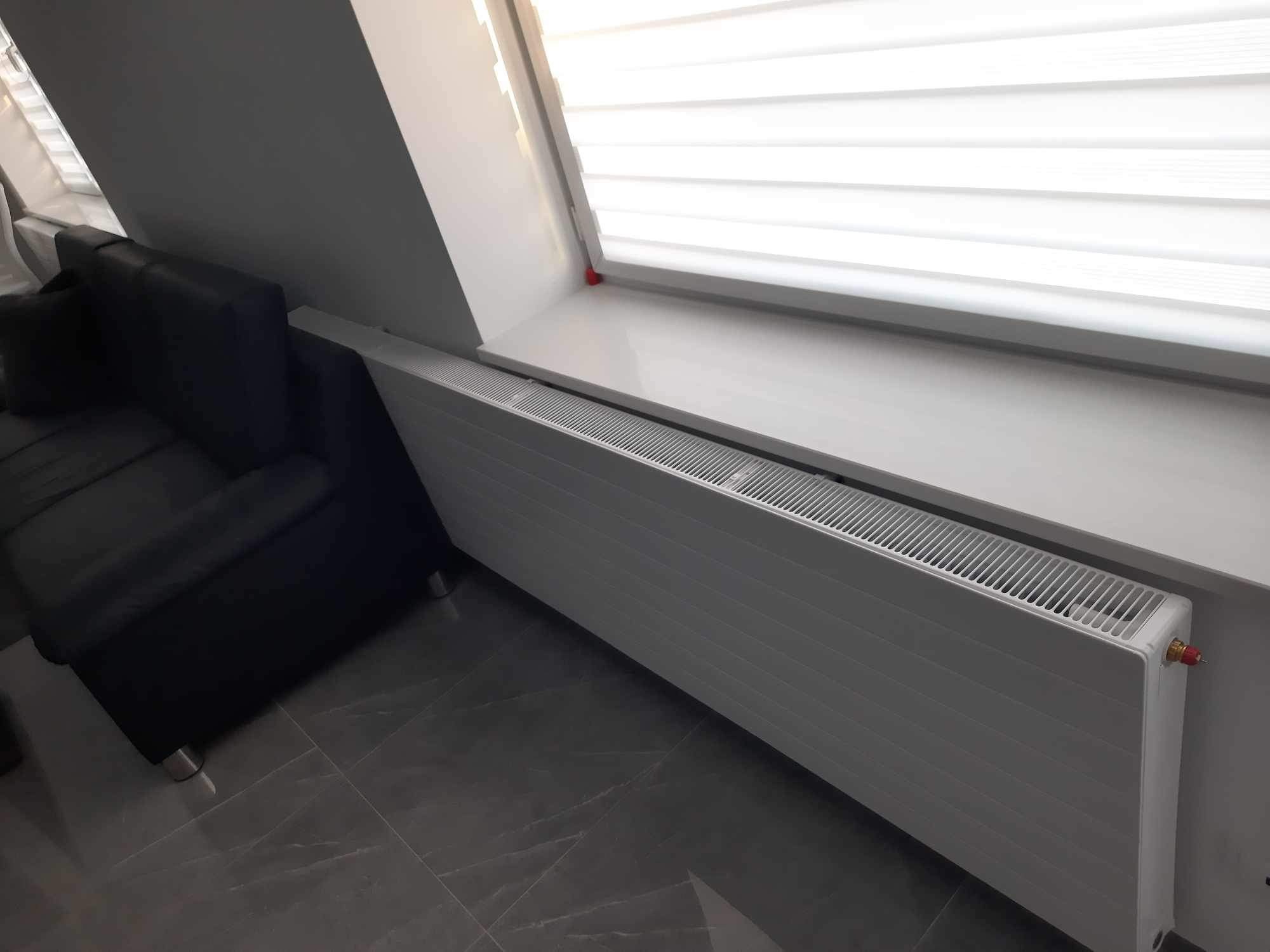 Biały grzejnik panelowy zamontowany pod oknem dachowym z białymi żaluzjami, obok ciemnoszarej sofy na posadzce z płytek imitujących marmur.