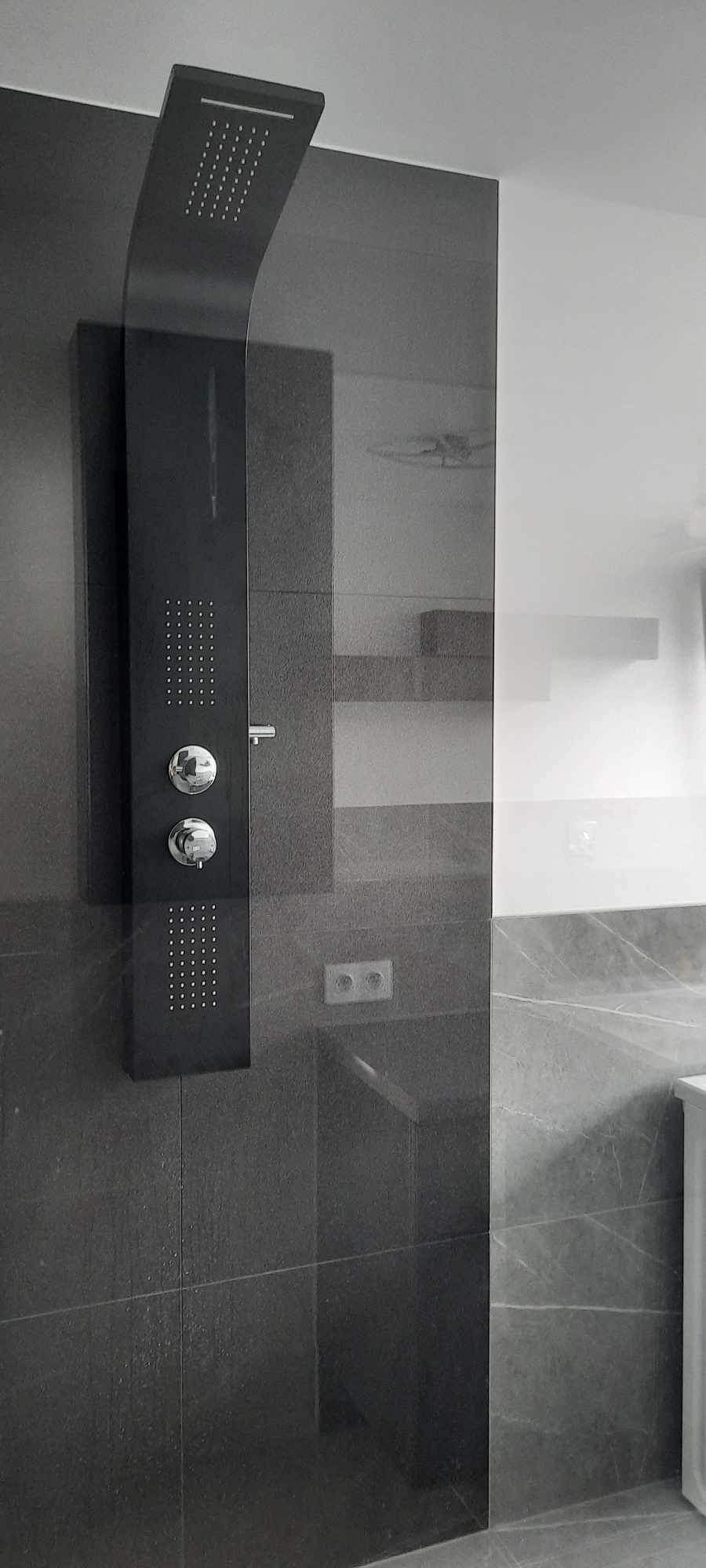 Nowoczesna kabina prysznicowa z czarnym panelem natryskowym na tle szarych płytek, widoczny fragment ściany z biało-szarymi płytkami imitującymi marmur.