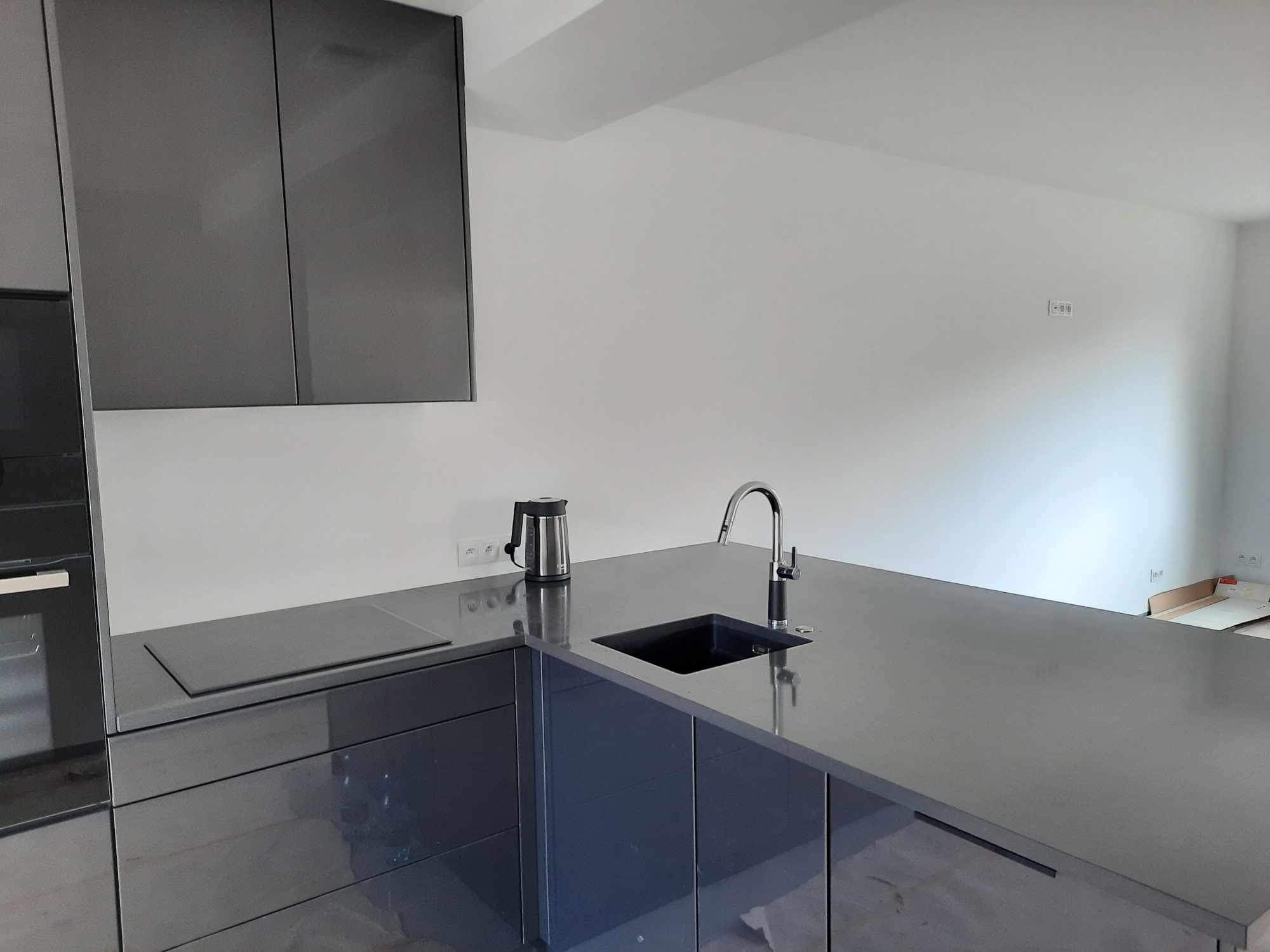 Nowoczesna, szara kuchnia z błyszczącymi frontami, minimalistycznym zlewem i czajnikiem na blacie, na tle białych ścian z widocznymi gniazdkami elektrycznymi.