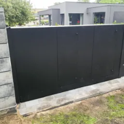 Nowoczesna, przesuwna brama wjazdowa w kolorze antracytowym z poziomymi akcentami, zamontowana na betonowym fundamencie z kostki brukowej, z widocznym fragmentem betonowego słupka ogrodzeniowego...