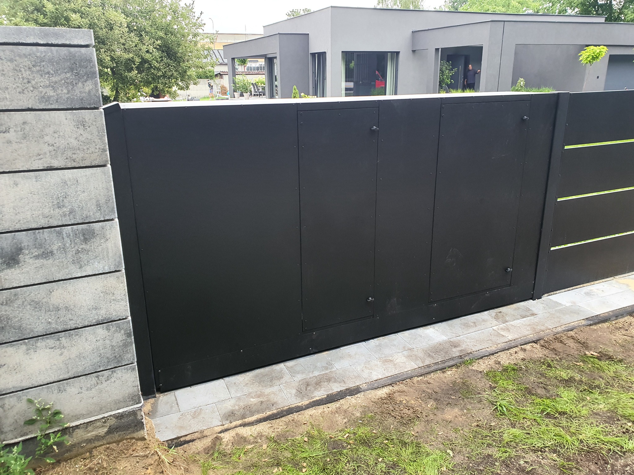 Nowoczesna, przesuwna brama wjazdowa w kolorze antracytowym z poziomymi akcentami, zamontowana na betonowym fundamencie z kostki brukowej, z widocznym fragmentem betonowego słupka ogrodzeniowego...