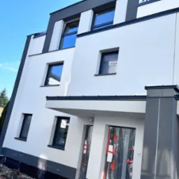 Nowoczesny budynek z białą elewacją i grafitowymi akcentami, zabezpieczone drzwi wejściowe folią malarską, widoczne fragmenty izolacji fundamentów i przygotowany teren wokół budynku.