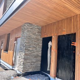 IVAN PRYVEDA - Nowoczesna elewacja domu w trakcie wykończenia: drewniana boazeria i kamień dekoracyjny. Widoczne zabezpieczenia okien i materiały budowlane.