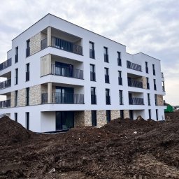 IVAN PRYVEDA - Nowoczesny, biały budynek wielorodzinny z balkonami i elementami kamiennej elewacji. W tle drugi budynek, na pierwszym planie ziemia i koparka.