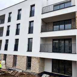 IVAN PRYVEDA - Nowoczesny budynek z białą elewacją i kamiennymi akcentami. Widoczni pracownicy w żółtych kamizelkach, teren budowy. Ujęcie z dołu, podkreślające architekturę.