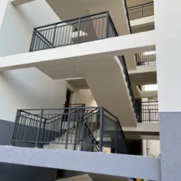 Wielopiętrowy korytarz z betonowymi schodami i metalowymi balustradami, świeżo pomalowany na biało i szaro, z widocznymi przewodami elektrycznymi zwisającymi z sufitu.