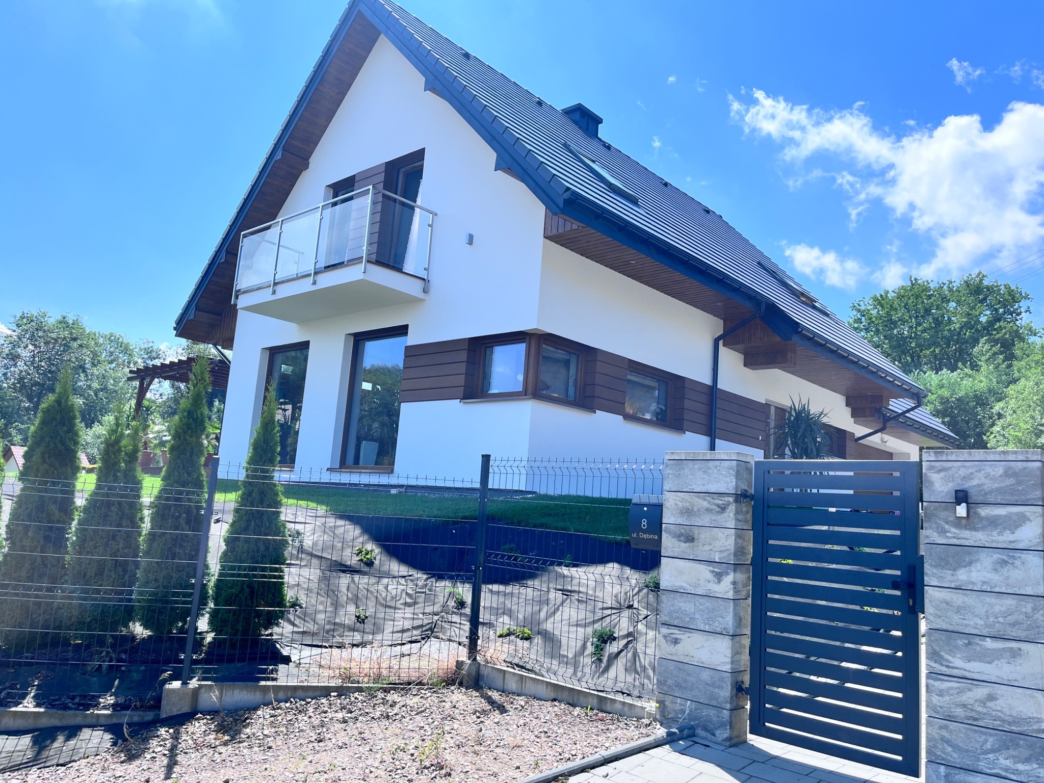 Nowoczesny dom jednorodzinny z białą elewacją, drewnianymi akcentami i balkonem ze szklaną balustradą, widok z niskiej perspektywy na tle błękitnego nieba z chmurami.