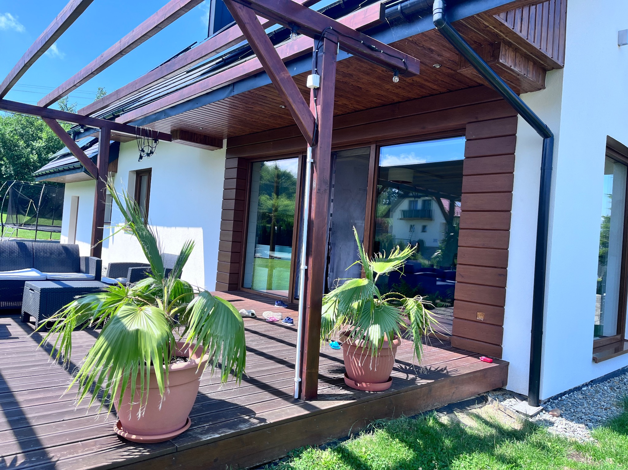 Nowoczesny dom z białymi ścianami i drewnianą elewacją, zadaszony taras z drewnianą pergolą, doniczkowe palmy i meble ogrodowe, widok z zewnątrz w słoneczny dzień.