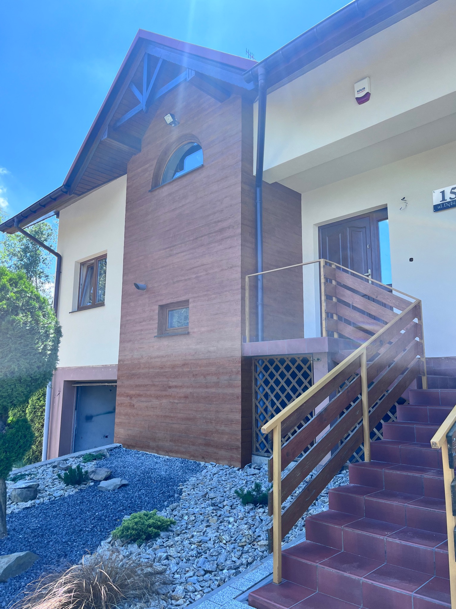 Dwukondygnacyjny dom z elewacją w odcieniach beżu i brązu, z drewnianymi elementami dekoracyjnymi i schodami wejściowymi z bordowymi płytkami, otoczony kamiennym ogrodem.