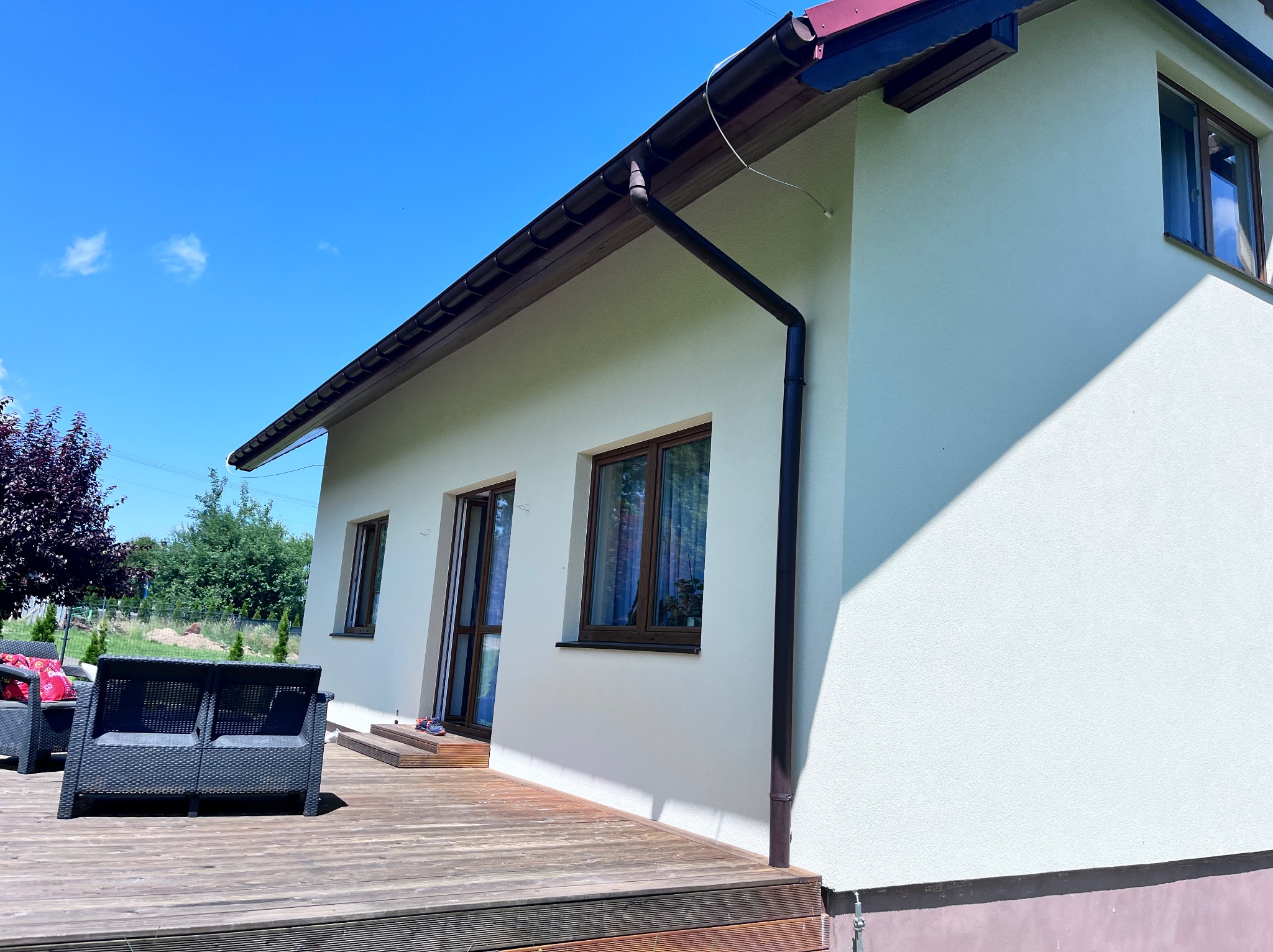 Jasny, otynkowany dom jednorodzinny w Krakowie z drewnianym tarasem i meblami ogrodowymi, uchwycony w słoneczny dzień.