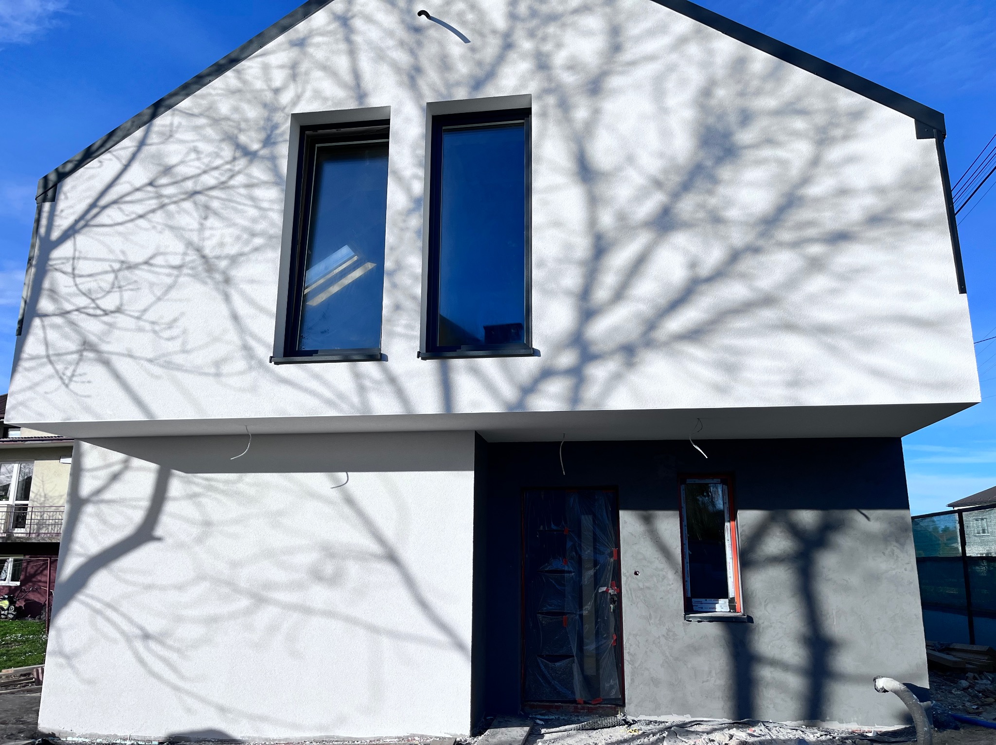 Dwukondygnacyjny dom z częściowo wykończoną elewacją: górna część biała, dolna szara, okna z czarnymi ramami, drzwi zabezpieczone folią, cień drzewa na ścianie.