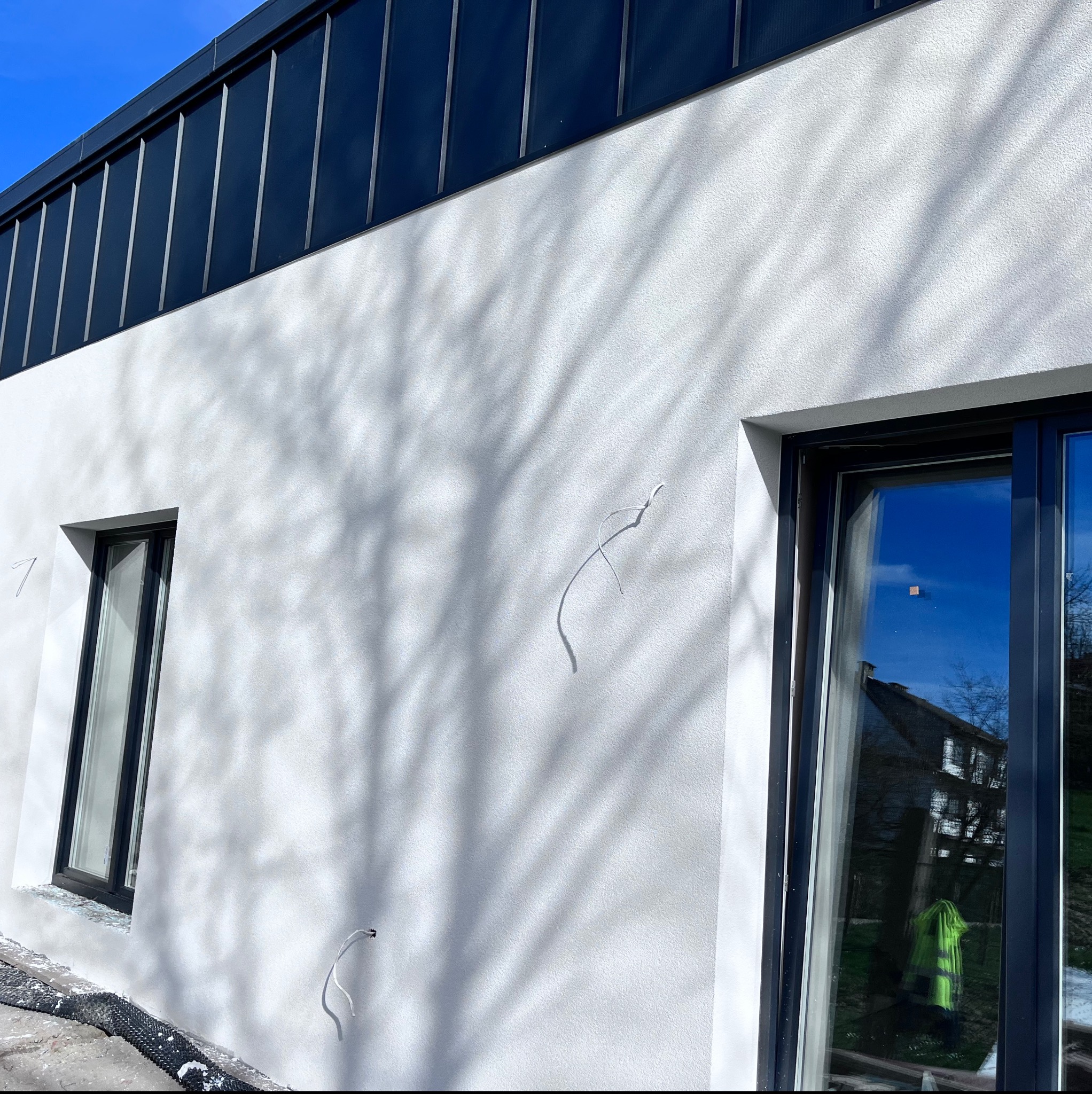 Jasna elewacja budynku z widocznymi przewodami elektrycznymi wystającymi ze ściany, cień drzewa na ścianie, czarne ramy okienne, widoczna siatka ochronna przy fundamencie.