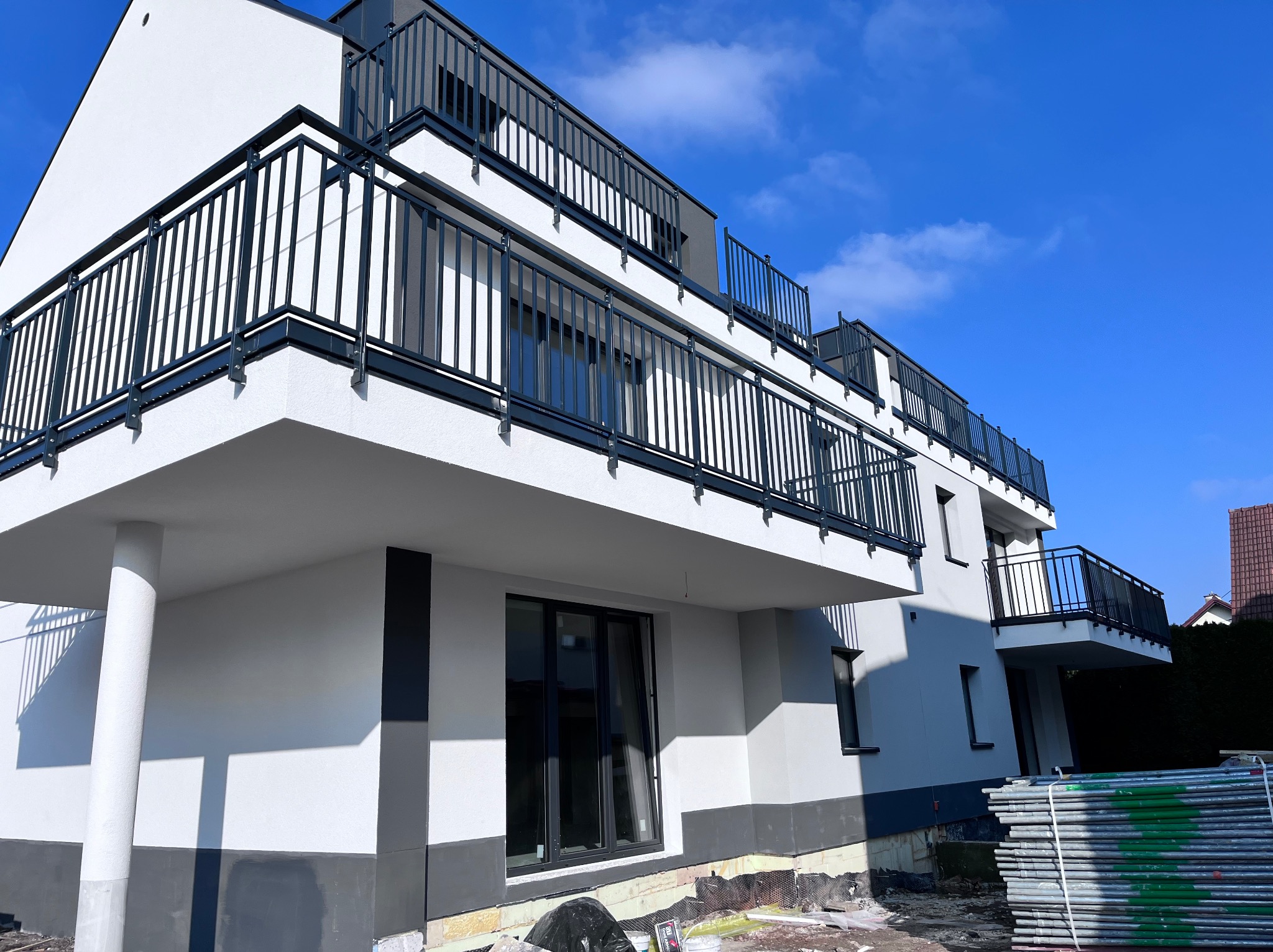 Nowoczesny budynek wielorodzinny z balkonami z czarną balustradą, świeżo otynkowany na biało i szaro, z widocznymi rusztowaniami i elementami przygotowania podłoża wokół budynku.