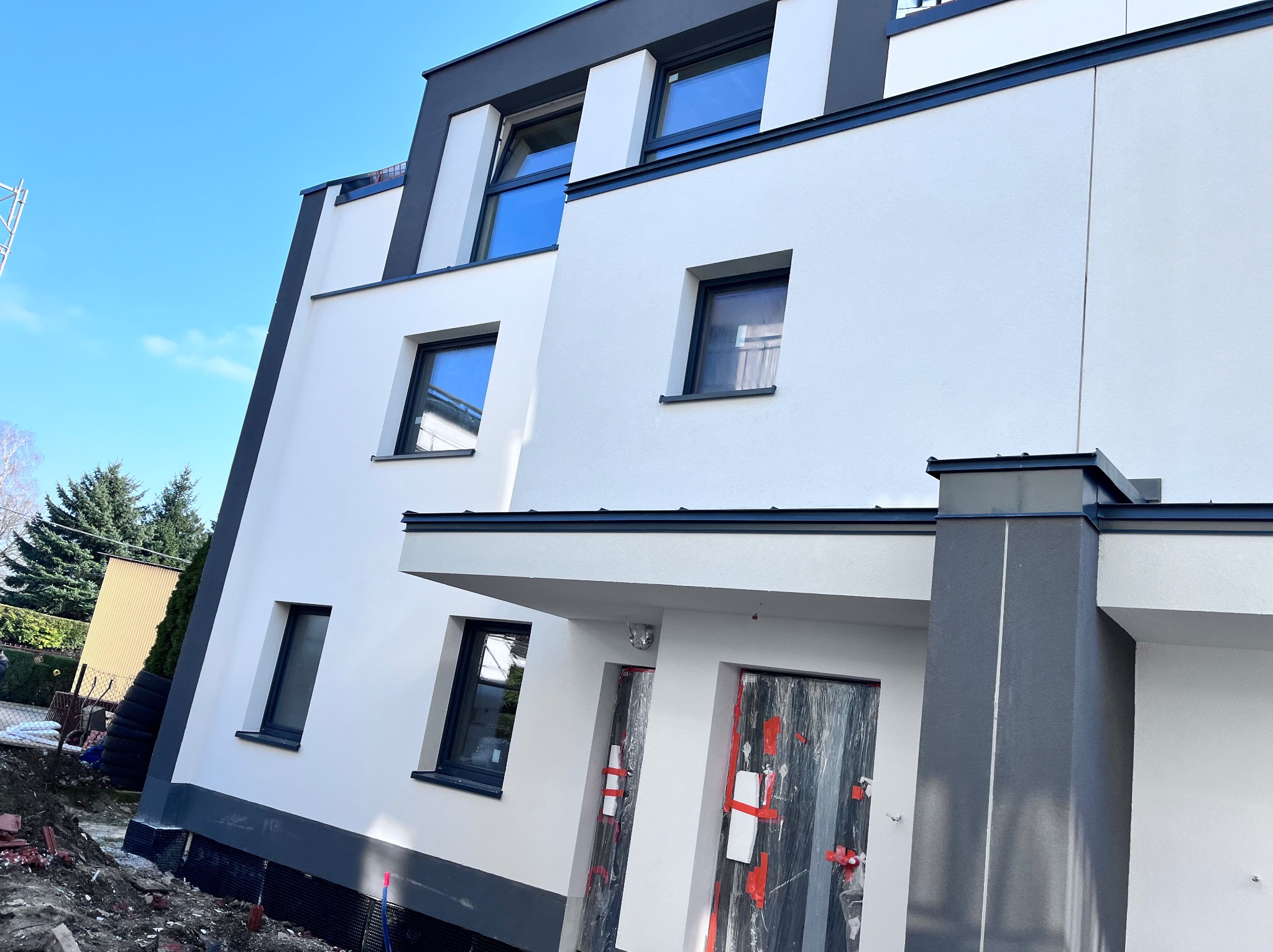 Nowoczesny budynek z białą elewacją i grafitowymi akcentami, zabezpieczone drzwi wejściowe folią malarską, widoczne fragmenty izolacji fundamentów i przygotowany teren wokół budynku.