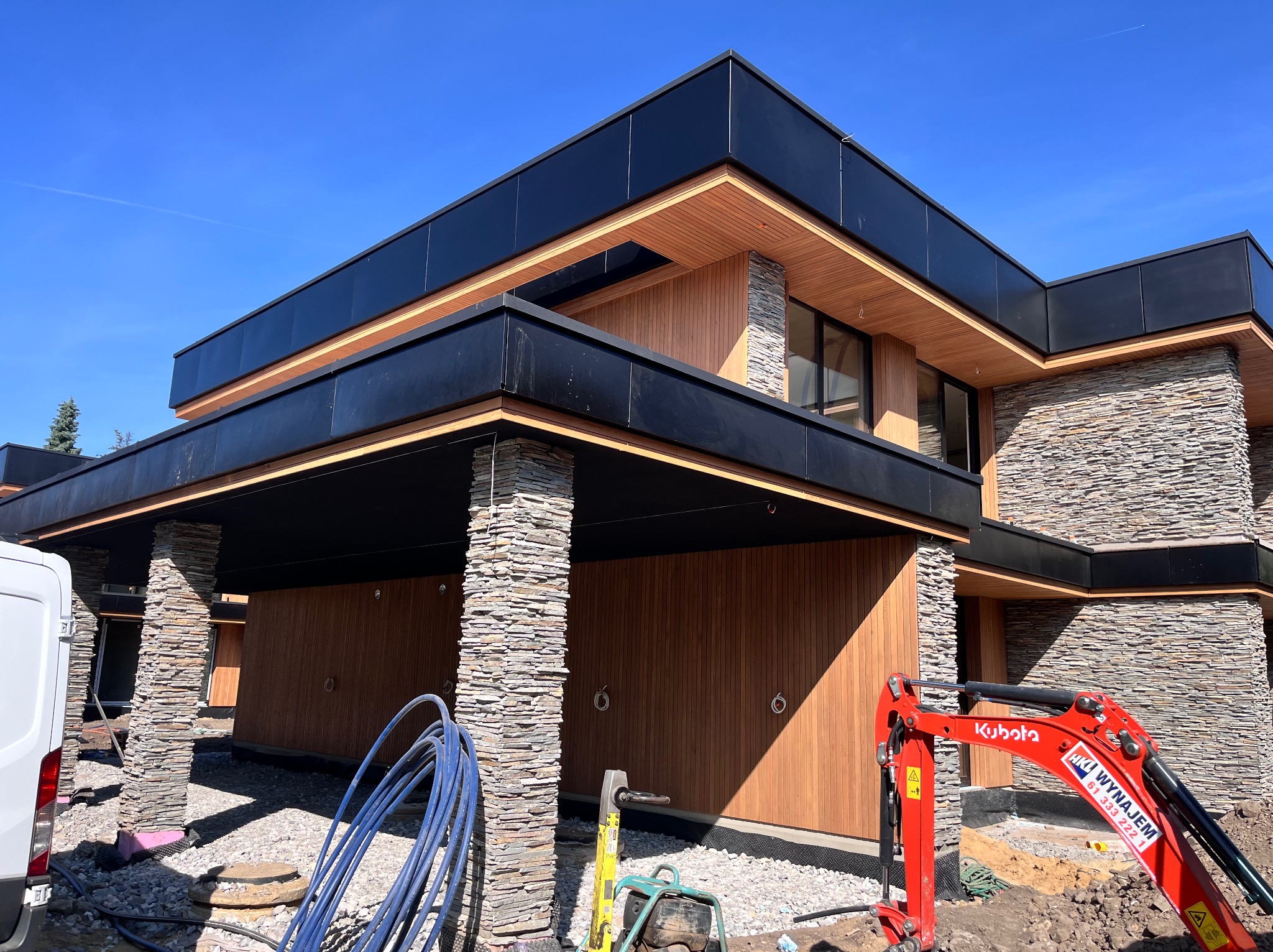 Nowoczesny dom z elewacją z kamienia i drewna, czarne elementy wykończeniowe, minikoparka Kubota na pierwszym planie, błękitne niebo w tle.