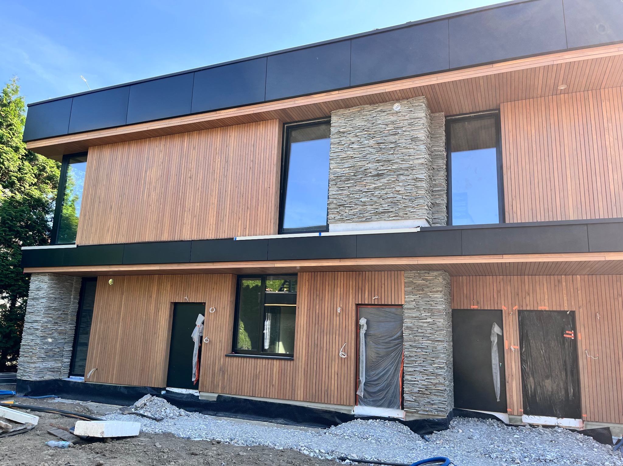 Nowoczesny dom w trakcie wykończenia elewacji: drewniana okładzina pionowa, kamienne filary, czarne elementy. Widoczne zabezpieczone drzwi i teren budowy.