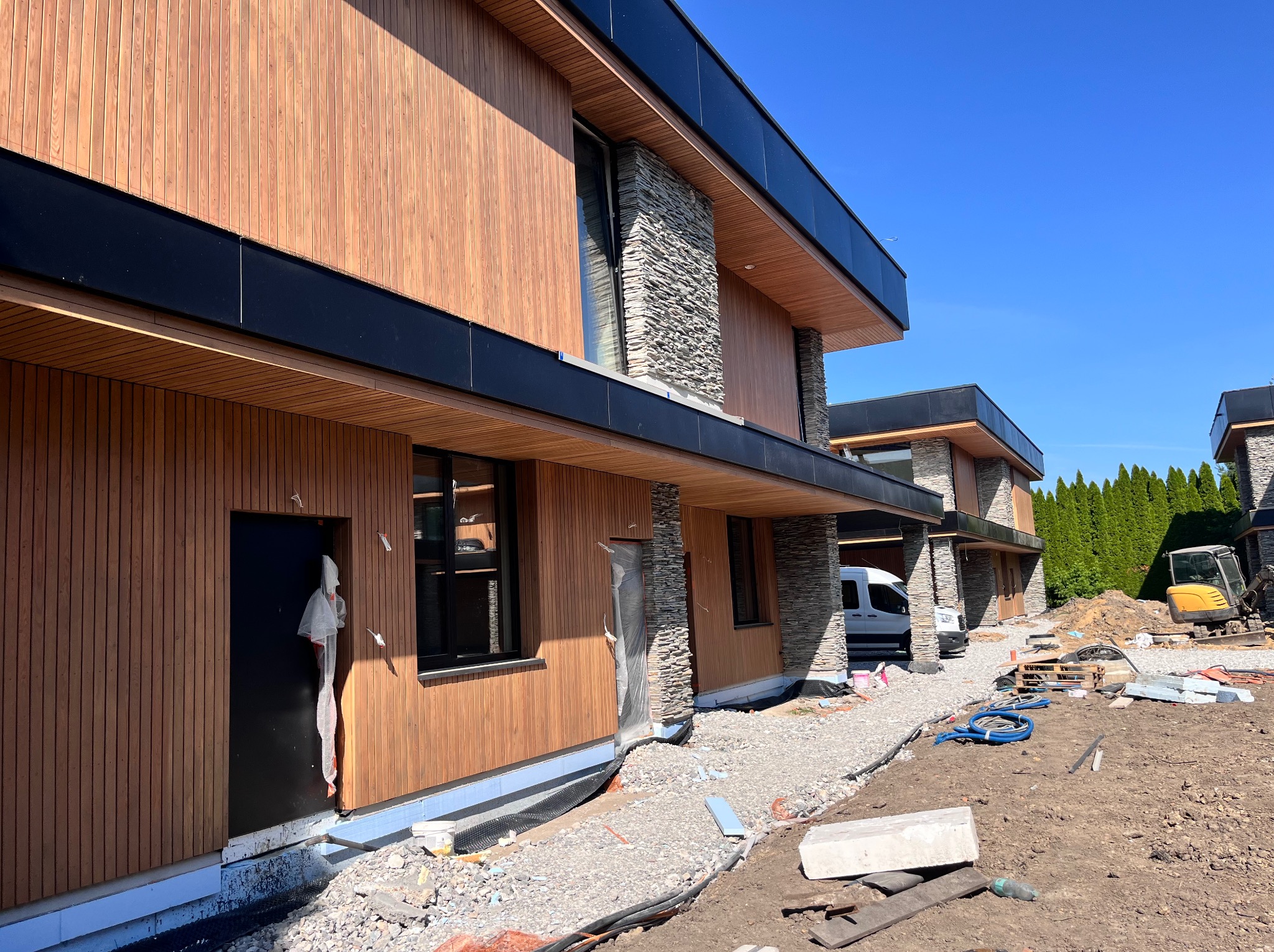 Nowoczesny dom z drewnianą elewacją i kamiennymi elementami wykończeniowymi, w trakcie prac wykończeniowych na zewnątrz budynku.