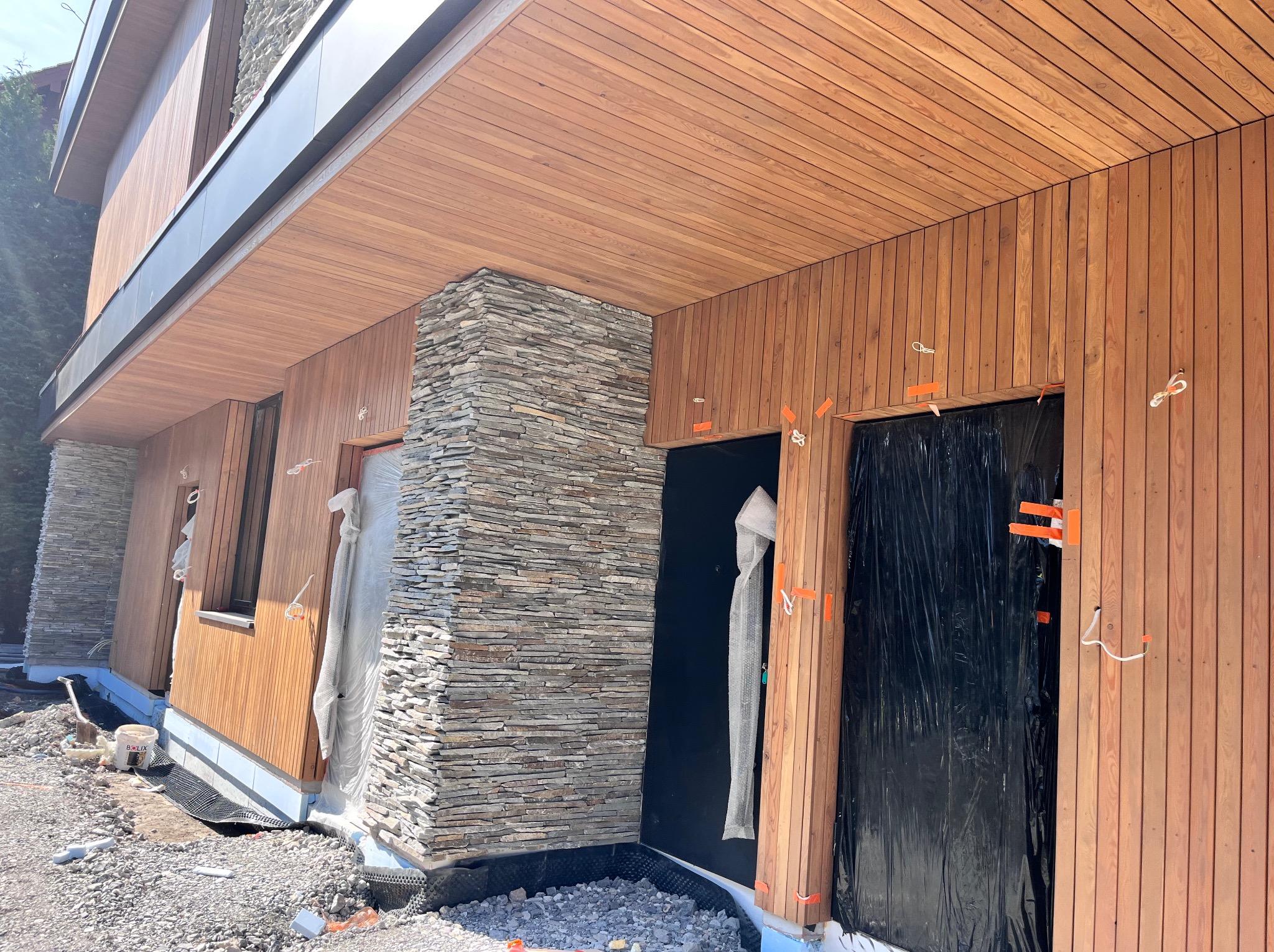 Nowoczesna elewacja domu w trakcie wykończenia: drewniana boazeria i kamień dekoracyjny. Widoczne zabezpieczenia okien i materiały budowlane.