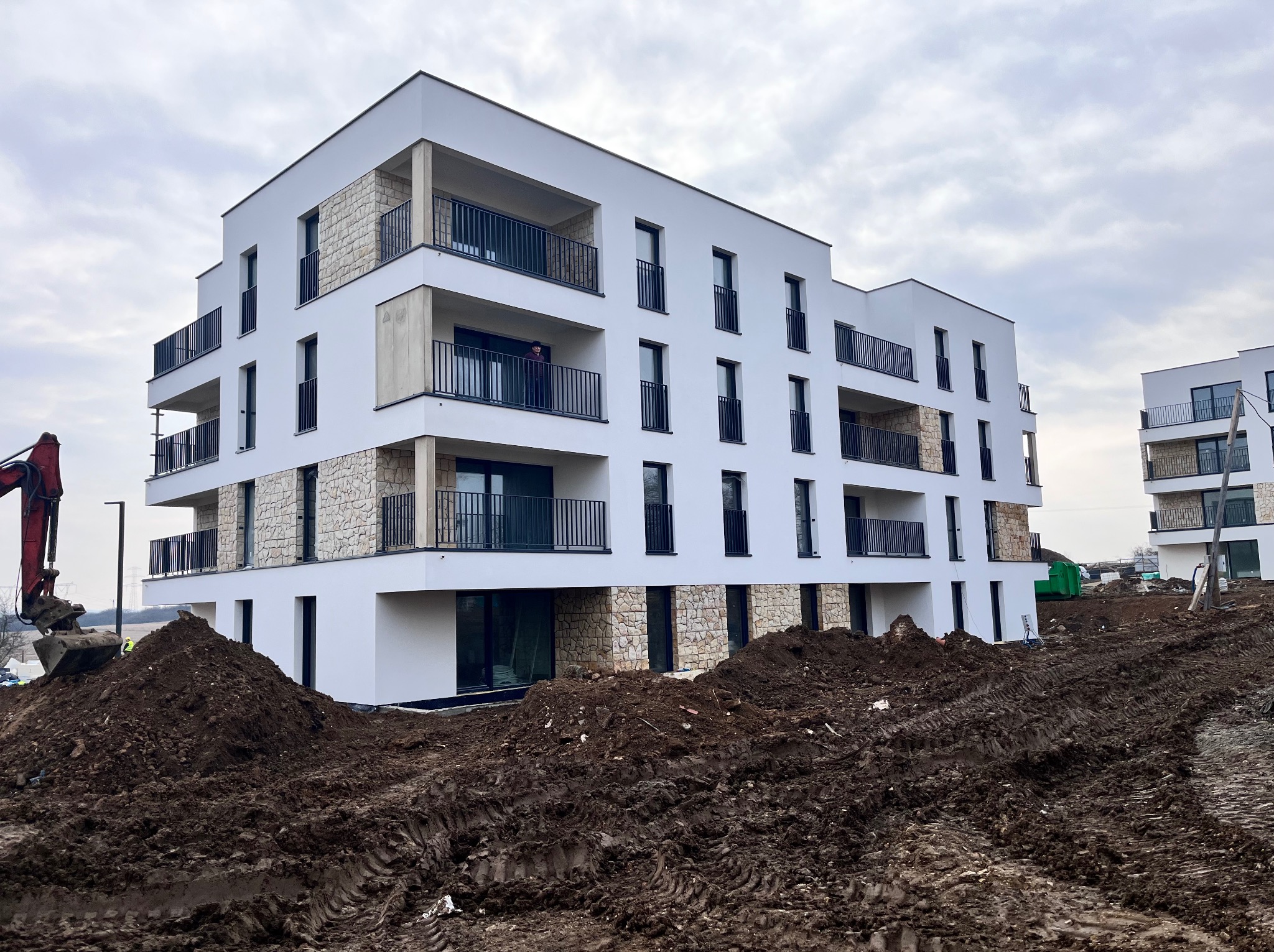 Nowoczesny, biały budynek wielorodzinny z balkonami i elementami kamiennej elewacji. W tle drugi budynek, na pierwszym planie ziemia i koparka.