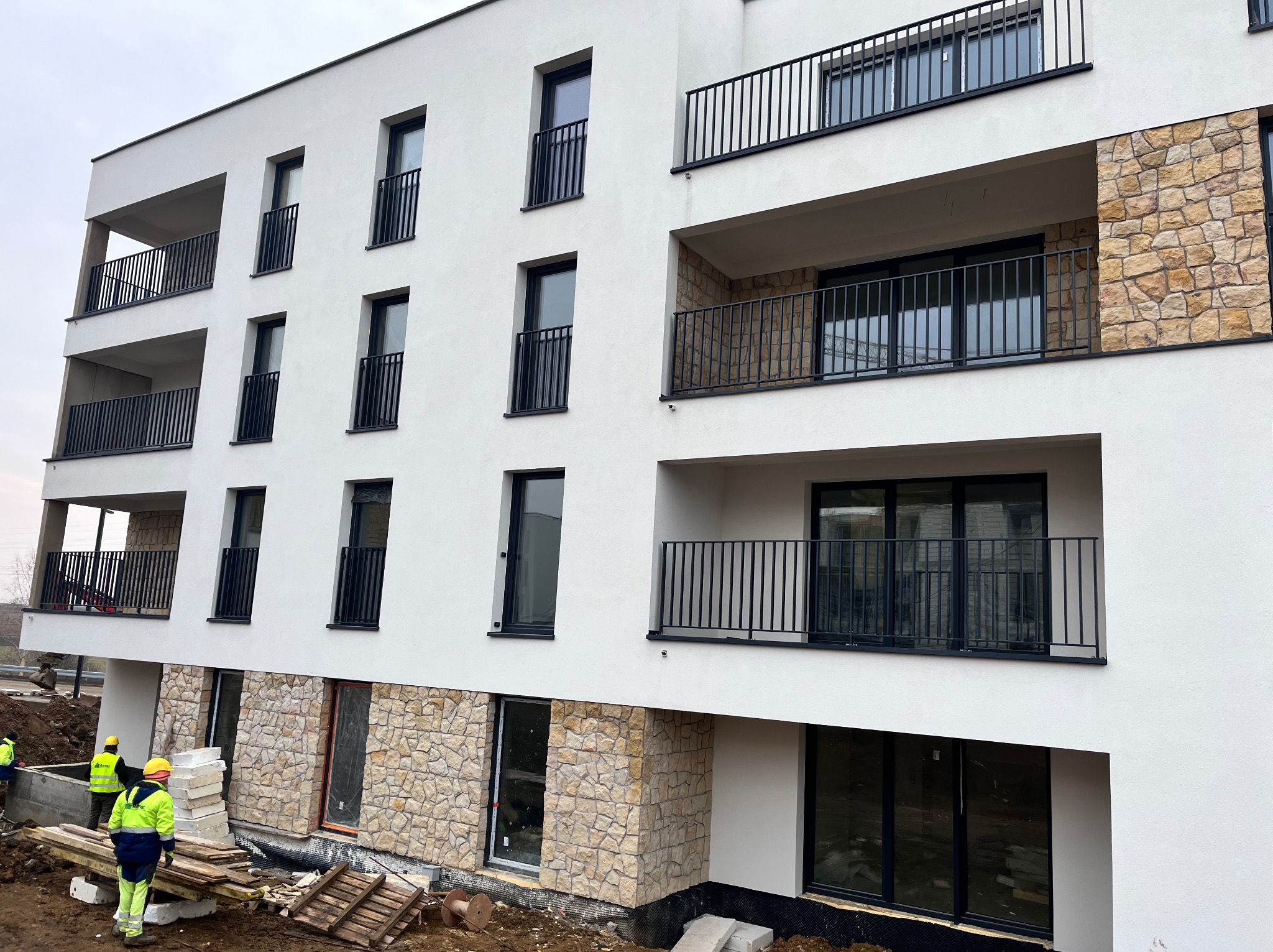 Nowoczesny budynek z białą elewacją i kamiennymi akcentami. Widoczni pracownicy w żółtych kamizelkach, teren budowy. Ujęcie z dołu, podkreślające architekturę.