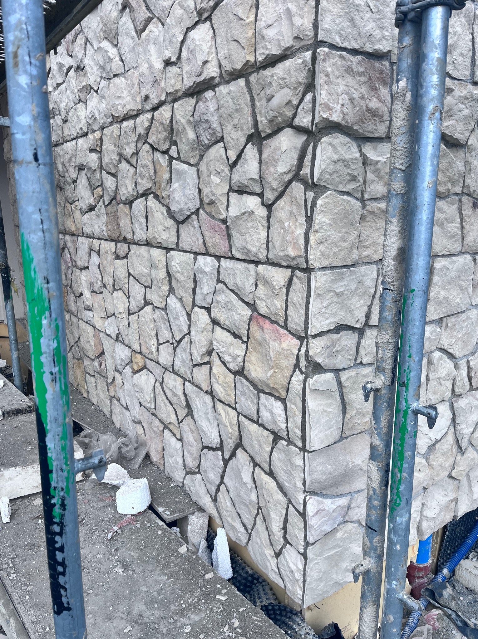 Wykończona elewacja budynku z kamienia naturalnego w odcieniach szarości i beżu. Widoczne rusztowania i fragmenty materiałów budowlanych.