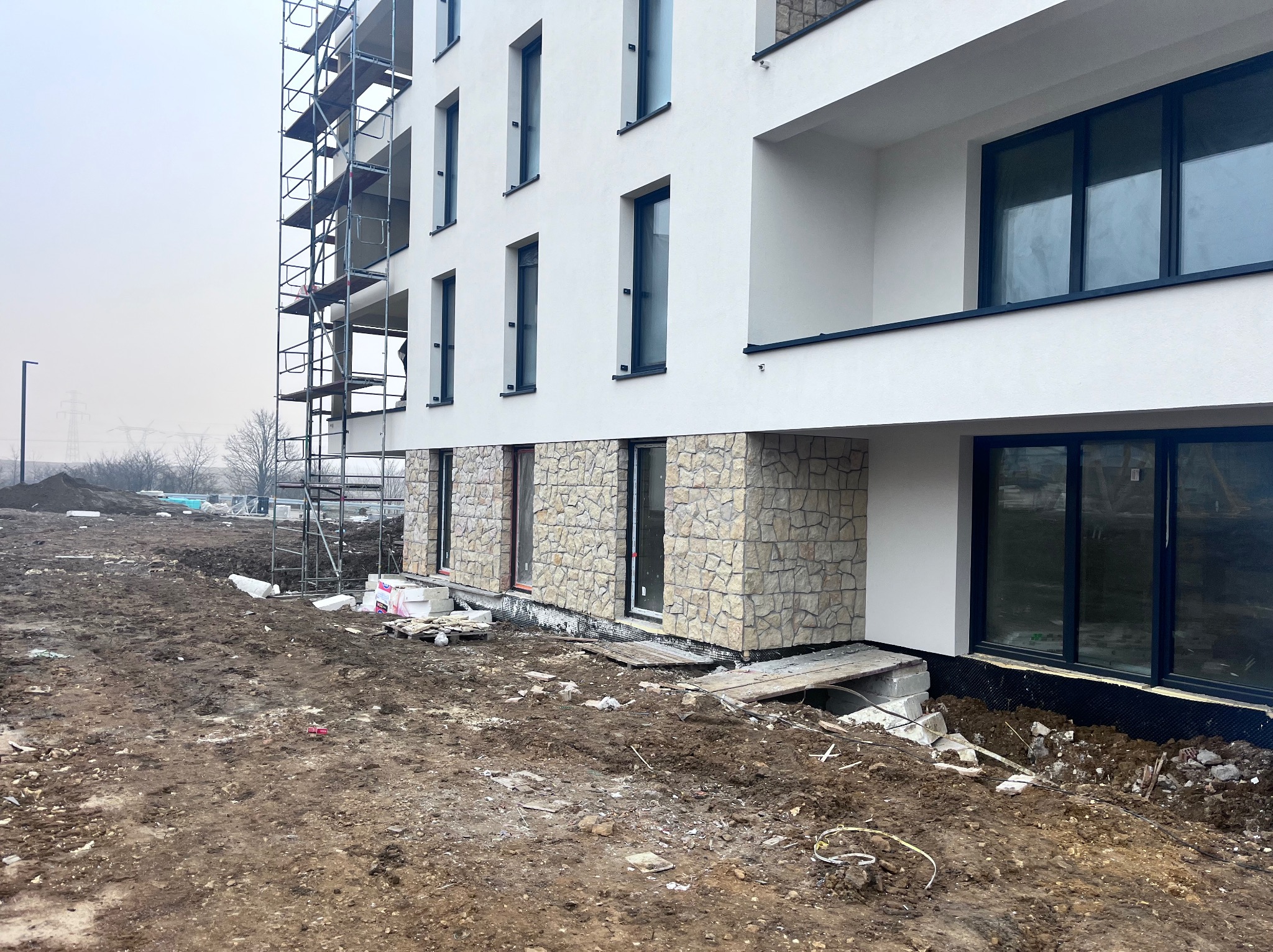 Nowoczesny budynek z białą elewacją i kamiennym wykończeniem dolnej części. Widoczne rusztowania i teren budowy. Okna w ciemnych ramach.
