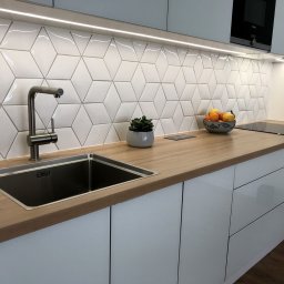 WeldJop - Nowoczesna kuchnia w Bydgoszczy: białe szafki, drewniany blat, geometryczne płytki na ścianie, minimalistyczny zlew i bateria. Oświetlenie LED pod szafkami.