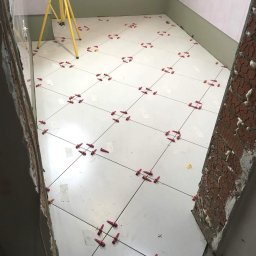 WeldJop - Białe płytki podłogowe w trakcie układania w łazience, z użyciem czerwonych krzyżyków dystansowych i poziomicy laserowej. Widoczne surowe ściany i logo firmy.