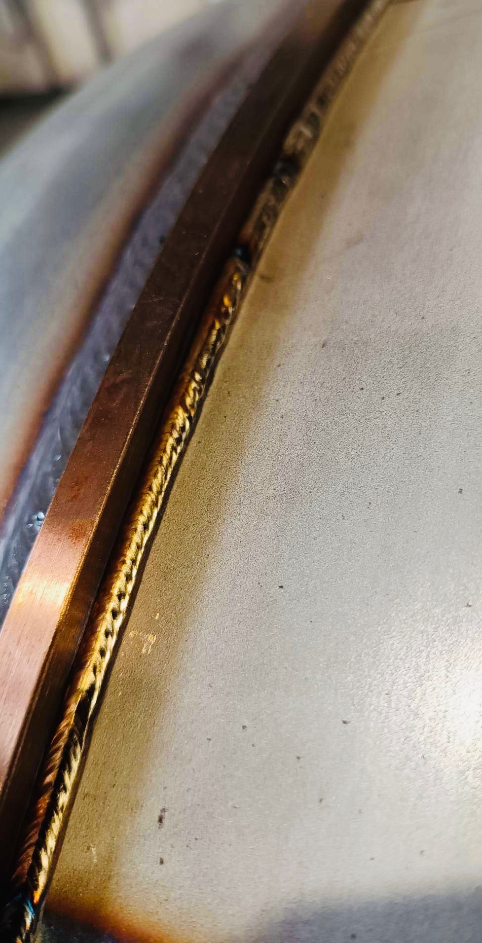 Szczegółowe zbliżenie na spawany szew na metalowej powierzchni z widocznymi przebarwieniami termicznymi i miedzianą listwą dociskową.